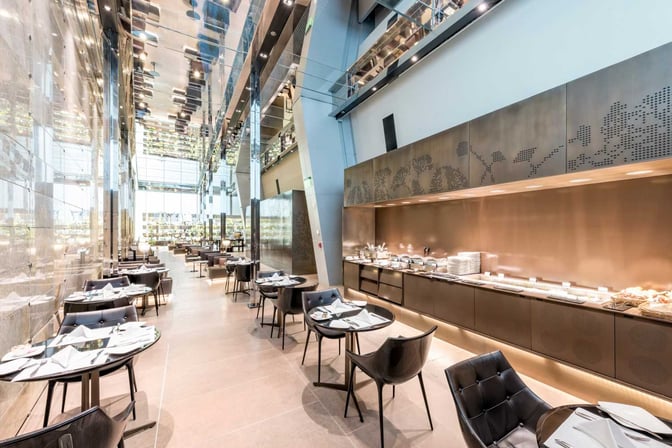 The Al Maha Lounge at Doha Hamad International Airport features a dining area with table seating and a buffet offering various dishes.