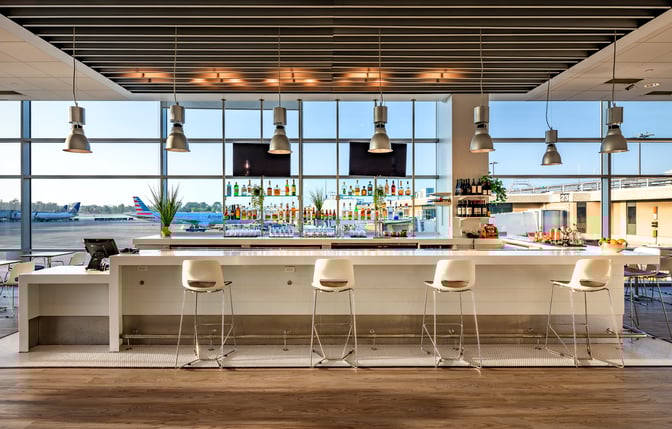 The Aspire Lounge at San Diego International Airport features a bar with high stools and a selection of spirits, overlooking the tarmac.