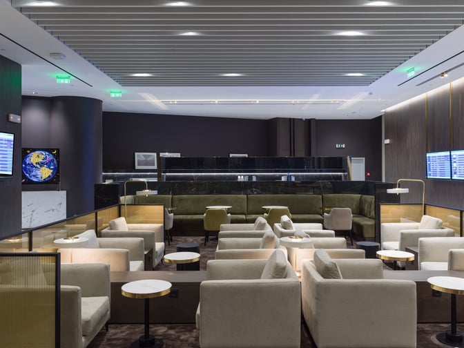 The seating area in Goldair Handling Lounge at Athens International Airport features comfortable chairs and tables with flight information screens visible.