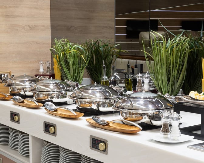 The buffet area at MERA Business Lounge Terminal 4 Cancun Airport offers self-serve chafing dishes with condiments and utensils.