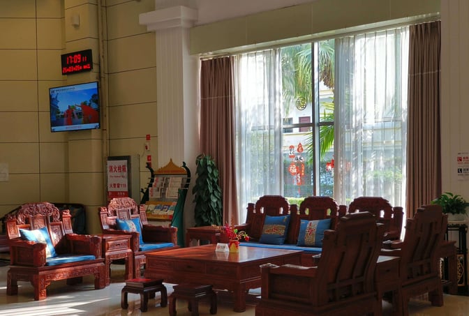The seating area in Dehong Mangshi Airport's VIP Lounge features intricately carved wooden chairs and a coffee table near large windows.