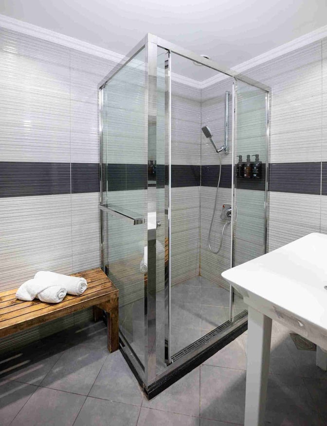 The shower facility at Sleepover Concourse D, Dubai International Airport, features a glass enclosure, wooden bench, and complimentary toiletries.