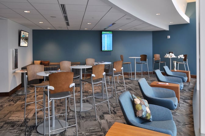 The seating area in The Club ATL features high-top tables and lounge chairs, with a departures screen for flight information.