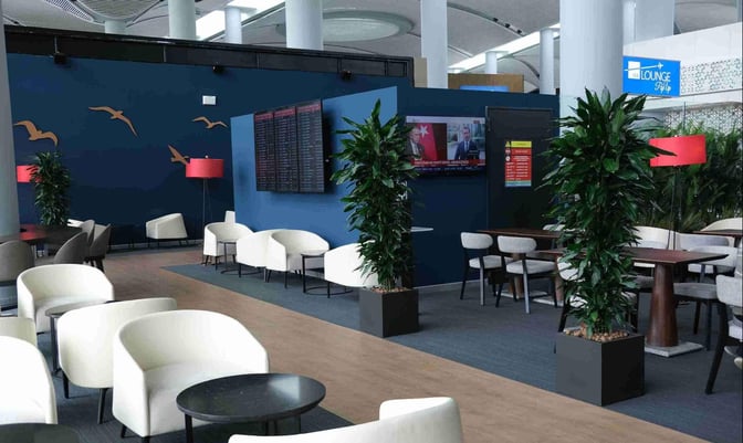 The IGA Pop Up Lounge at Istanbul Airport features comfortable seating areas with tables, flight information screens, and decorative plants.