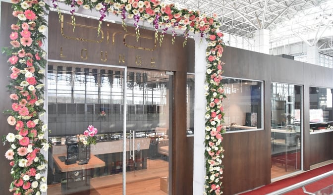 The Bird Lounge entrance at Jolly Grant Airport features floral decorations and glass doors, located in the terminal area.