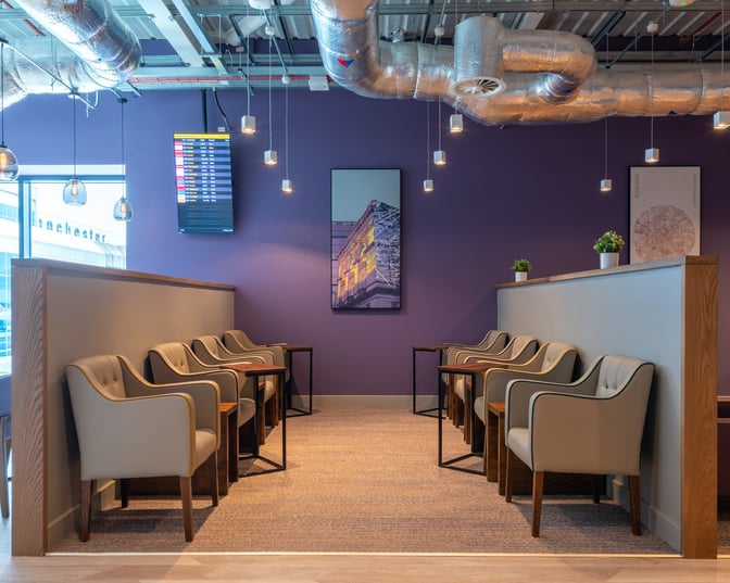 Seating area in Aspire Lounge at Manchester Airport features comfortable chairs and small tables, with a flight information screen visible.