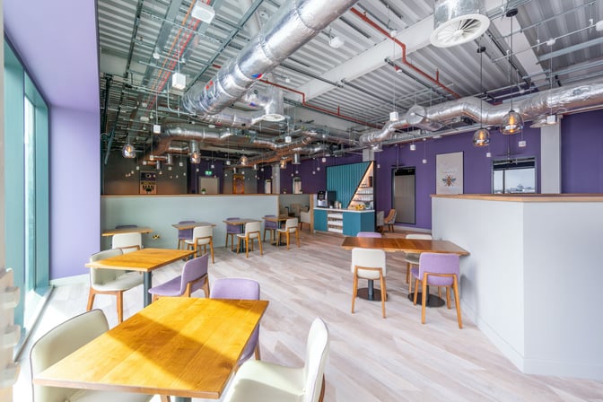 The Aspire Lounge at Manchester Airport features a seating area with tables and chairs, adjacent to a self-serve beverage station.