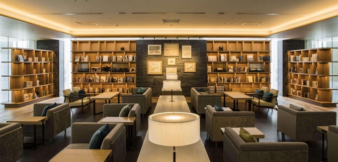 The seating area at CAFÉ SKY LIBRARY in New Chitose Airport features comfortable chairs and tables surrounded by bookshelves.