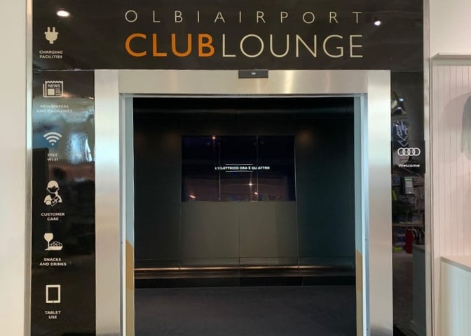 The entrance to the Olbia Airport Club Lounge features signage highlighting amenities like charging facilities, free WiFi, and snacks and drinks.