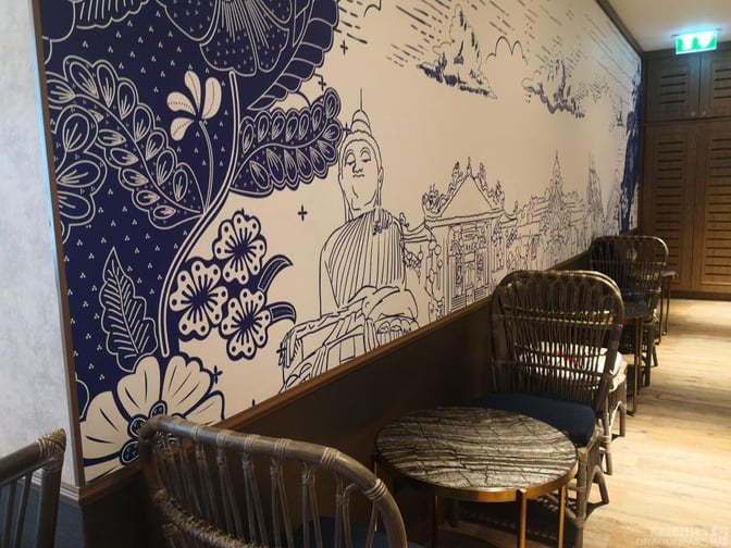 Seating area in Bangkok Airways Blue Ribbon Lounge at Phuket International Airport features wicker chairs and tables alongside a decorative wall mural.