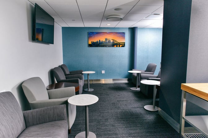 The seating area in The Club PIT at Pittsburgh International Airport features armchairs and small tables, with a wall-mounted TV.