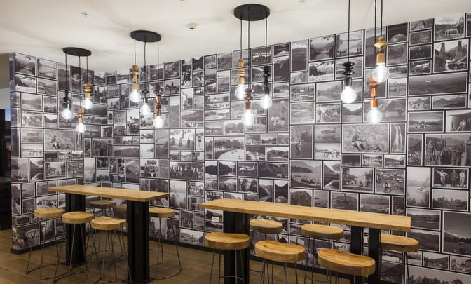High-top tables with stools are set against a photo collage wall in the Manaia Lounge at Queenstown Airport.