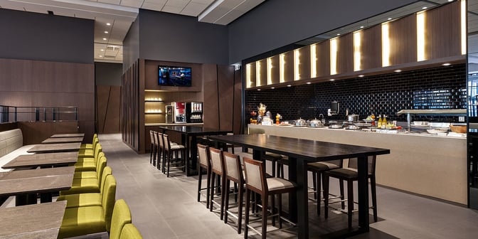 The Plaza Premium Lounge at Vancouver International Airport features a self-serve food area with high tables and seating near Domestic Gate C29.