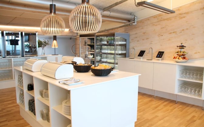 The Aalborg Airport Lounge features a self-serve food area with covered dishes, glassware, and a selection of fresh fruits.