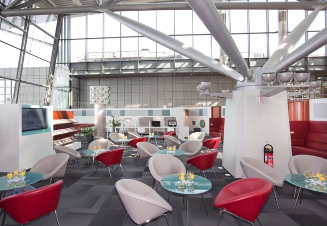 The seating area in Hamburg Airport's lounge features round tables with chairs and a view of the terminal's large windows.