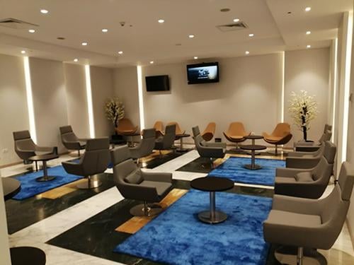 The seating area in Pearl Lounge at Alexandria International Airport features swivel chairs and small tables, with multiple TV screens for entertainment.