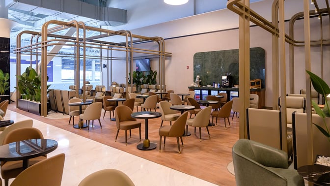 The seating area in the Extime Business Lounge at Almaty International Airport features tables and chairs with a nearby refreshment counter.