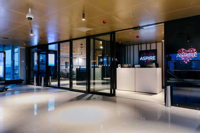 The entrance to the Aspire Lounge at Amsterdam Schiphol Airport features a reception desk and glass doors near Gate 26.