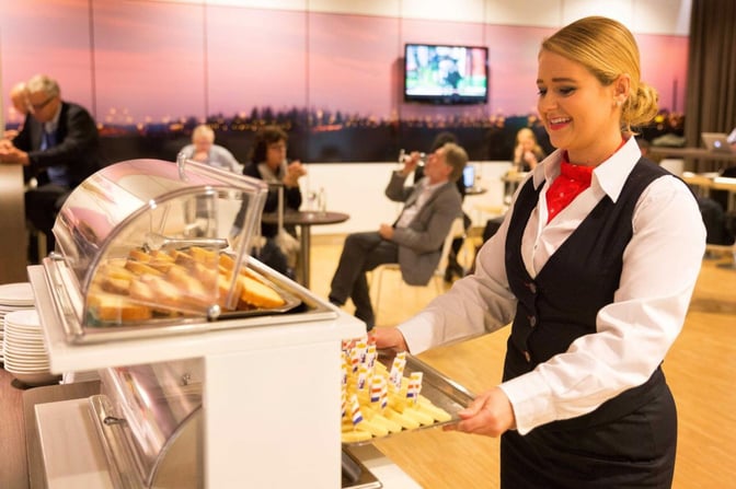 A staff member in the Aspire Lounge at Amsterdam Schiphol Airport serves food from a buffet area near Gate 26.