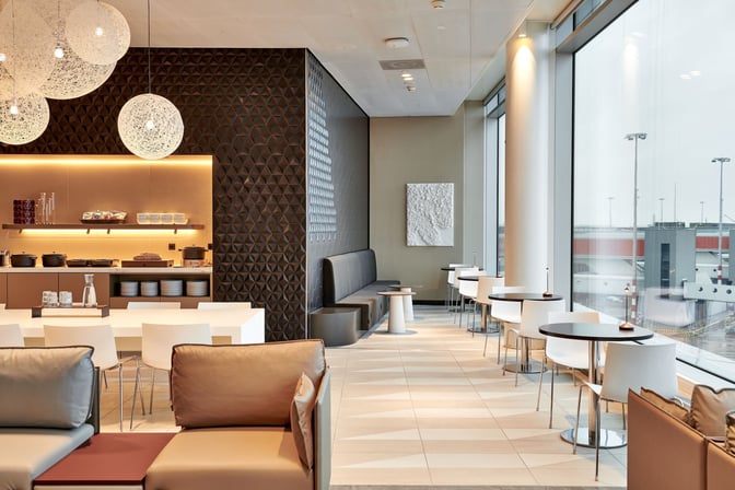 The Aspire Lounge at Amsterdam Schiphol Airport features a seating area with tables and chairs overlooking the airport tarmac.
