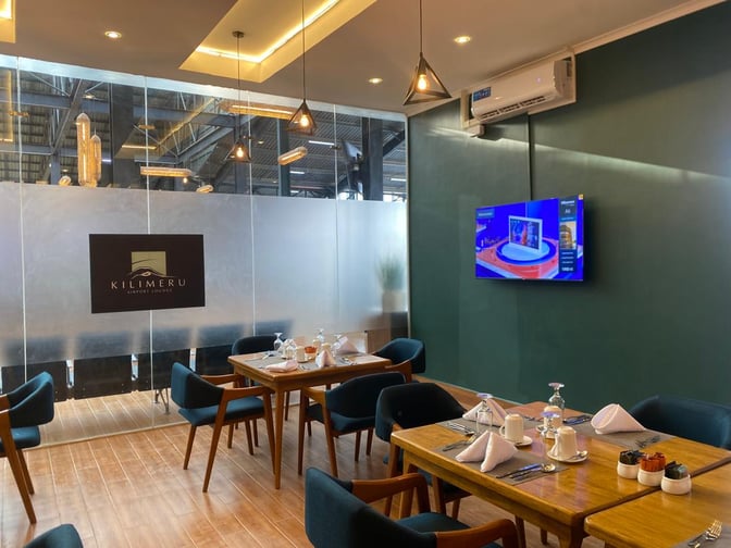 The Kilimeru Lounge at Arusha Airport features a dining area with set tables and a wall-mounted television for entertainment.