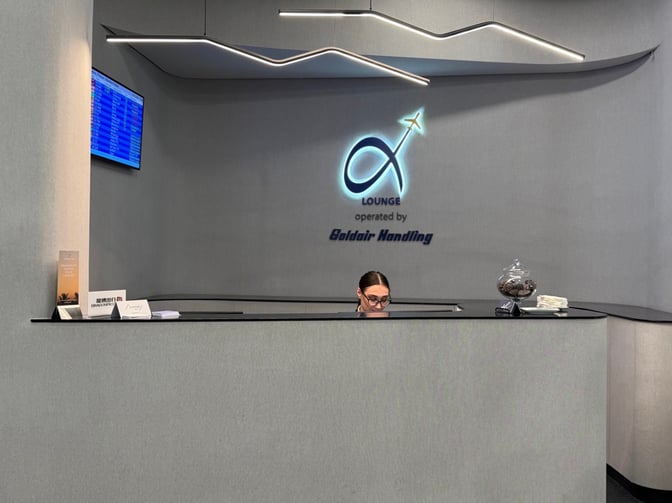 The reception desk at the Goldair Handling Lounge in Athens International Airport features a flight information screen and guest check-in materials.