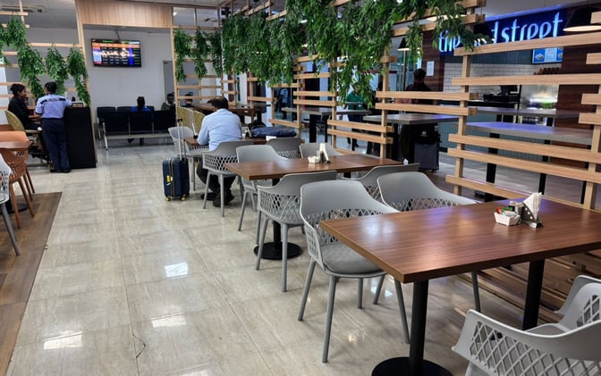 The seating area at Bagdogra Airport's Take Off Bar and Lounge features tables and chairs with nearby flight information screens.