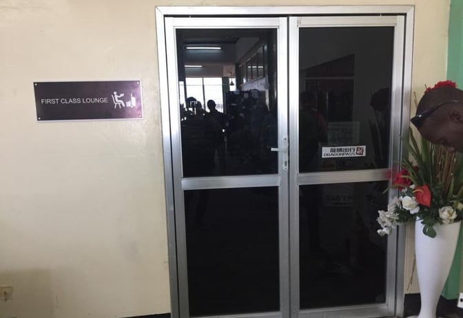 The entrance to the First Class Lounge at Banjul International Airport features a glass door and a sign indicating its location.