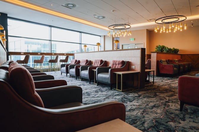 The Aspire Lounge at Belfast City Airport features comfortable seating with tables and a view of the airport through large windows.