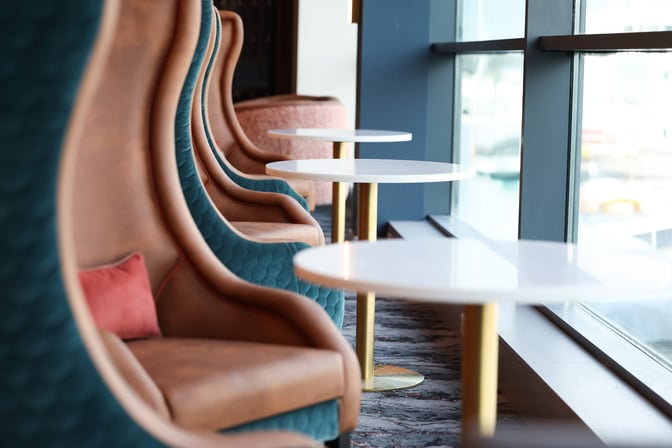 High-backed seating with small round tables is arranged along a window, offering views of the airport at Belfast City.