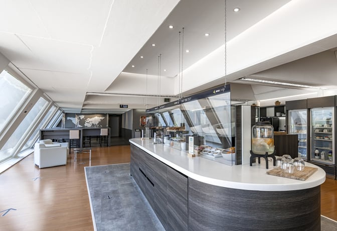 The Nervión VIP Lounge at Bilbao Airport features a self-serve food station with snacks and beverages, alongside seating areas with tables.