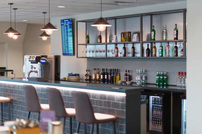 The Aspire Lounge South at Birmingham Airport features a bar with a selection of spirits, wines, and a digital departure board.