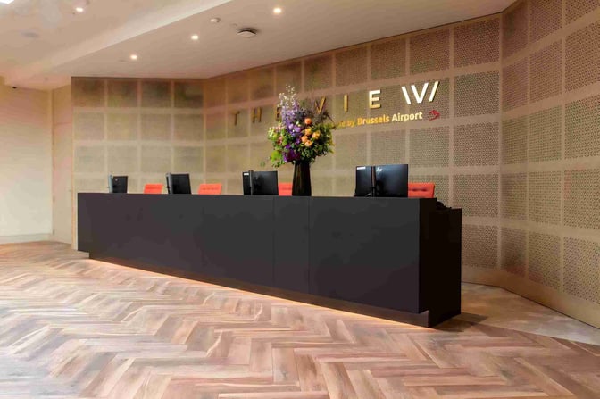 The reception area of The View at Brussels Airport features a long counter with computers and a decorative flower arrangement.
