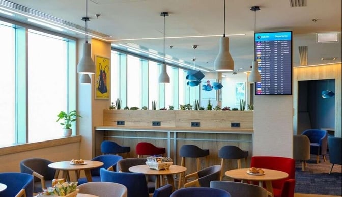 The seating area in Plaza Premium Lounge at Budapest Airport features tables with snacks and a flight information display.