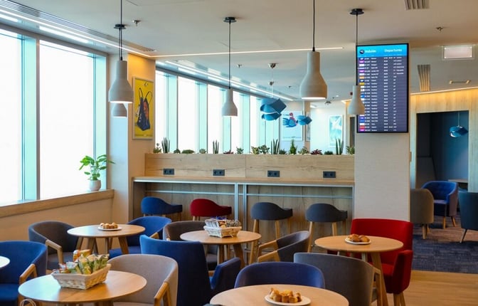Seating area in the Plaza Premium Lounge at Budapest Airport features round tables with snacks and a flight information screen.