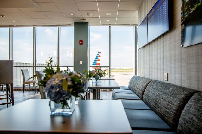 Seating area at The Club BUF features tables with floral arrangements and offers views of the airport runway through large windows.