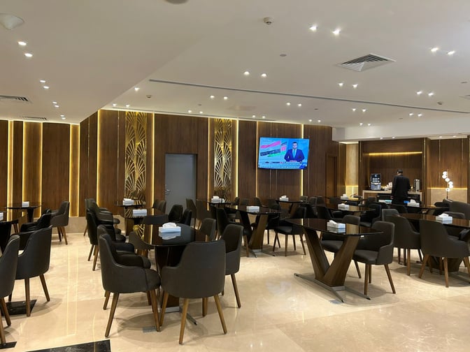 The CAC Lounge at Cairo International Airport Terminal 3E features a spacious seating area with tables and chairs, and a television for entertainment.