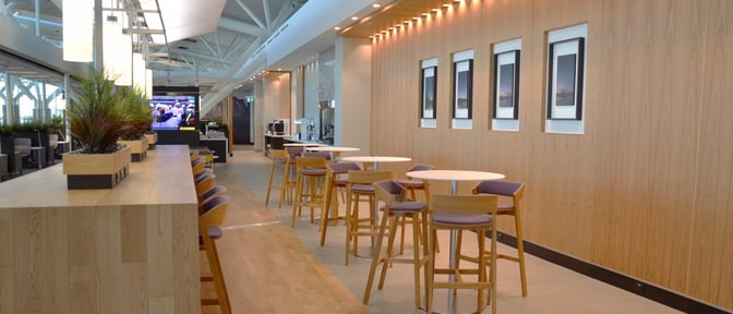 The Aspire Lounge at Calgary International Airport features a seating area with high tables and chairs, adjacent to a self-serve beverage station.