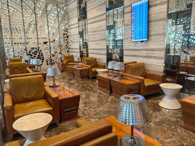 The seating area at Adani East Lounge in Chhatrapati Shivaji International Airport features comfortable chairs, side tables, and a flight information display.