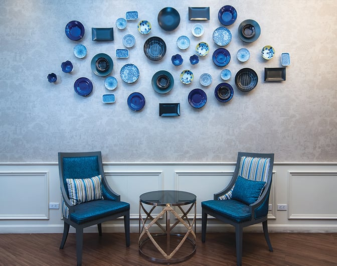 Two chairs and a small table are arranged in a seating area beneath decorative wall art at the Bangkok Airways Blue Ribbon Lounge.