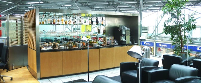 The Lounge Koeln-Bonn at Cologne Bonn Airport features a self-serve bar with spirits, glassware, and coffee machines, adjacent to seating areas.