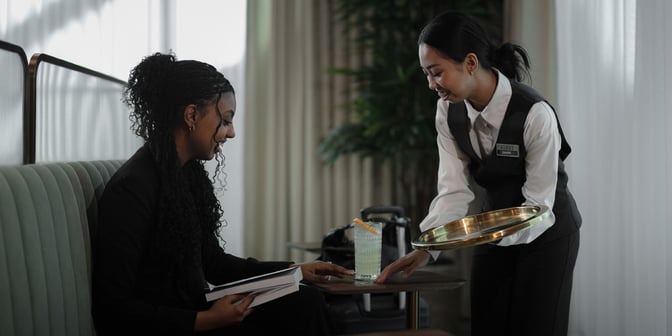 A server at Plaza Premium First Lounge, Dallas-Fort Worth, delivers a drink to a guest seated at a table.