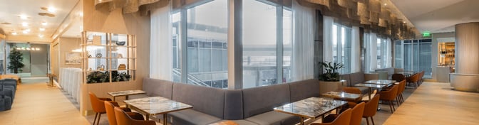 The seating area in the Plaza Premium Lounge at Dallas-Fort Worth International Airport features tables with booth and chair options near large windows.