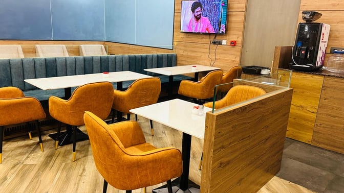 The seating area at Airr Lounge features tables with cushioned chairs and a TV, adjacent to a self-serve coffee station.