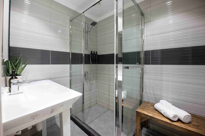 The shower area in Sleepover Concourse D at Dubai International Airport features a glass-enclosed shower, sink, and wooden bench with towels.