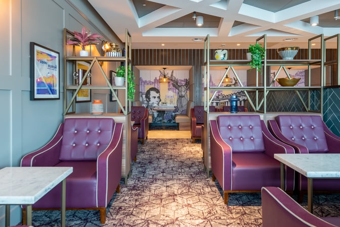 Seating area in Aspire Lounge, Gate 16 at Edinburgh Airport, features cushioned chairs and tables with decorative shelving and artwork.