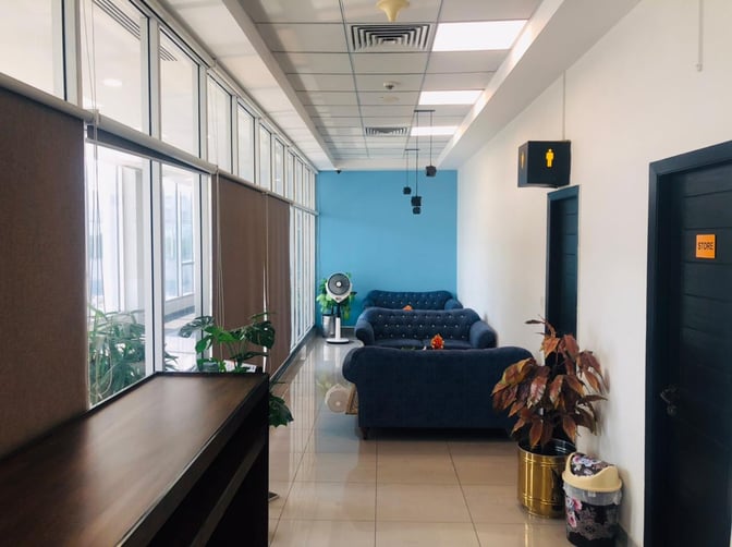 The seating area in Prestigio Lounge at Faisalabad International Airport features comfortable sofas and a fan, with nearby restroom access.