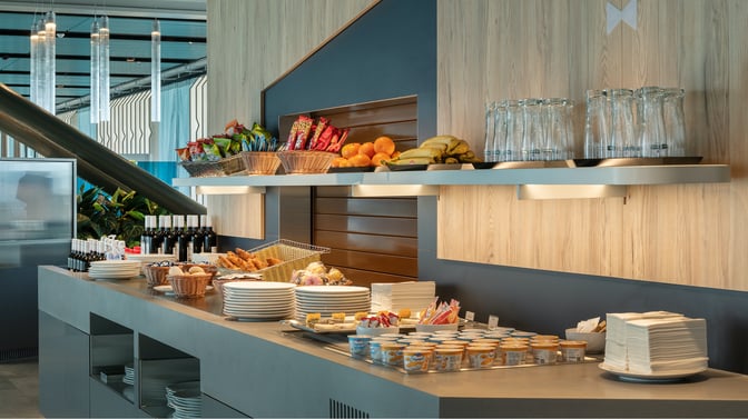Self-serve food station at Crystal Lounge, Geneva Airport, offers snacks, fruits, pastries, yogurt, and beverages for travelers.