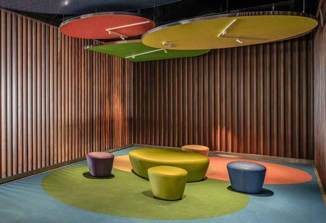 The seating area in Sala Galdós at Gran Canaria Airport features colorful, cushioned stools and benches with overhead circular panels.