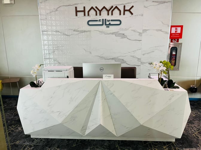 The reception desk at Hayyak Lounge in Bisha Domestic Airport features a geometric design with a computer and printer setup.